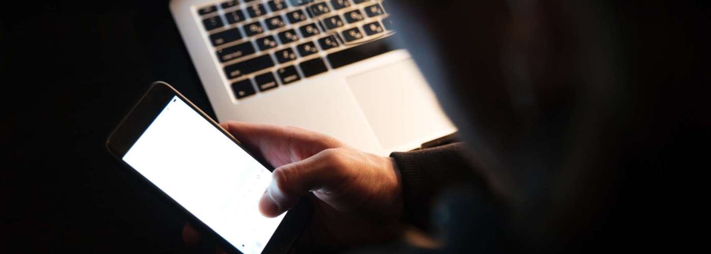 Cropped photo of young concentrated man using laptop computer and phone at home indoors at night.